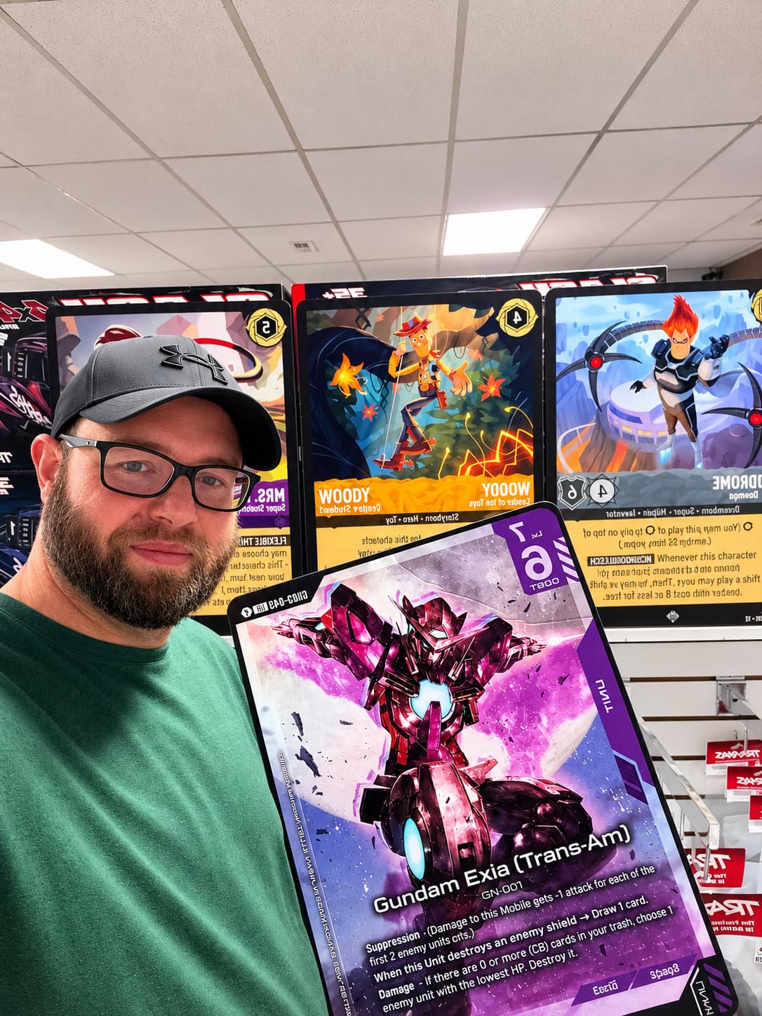 Bearded man holding a large Gundam Exia trading card with Woody and Syndrome cards behind.
