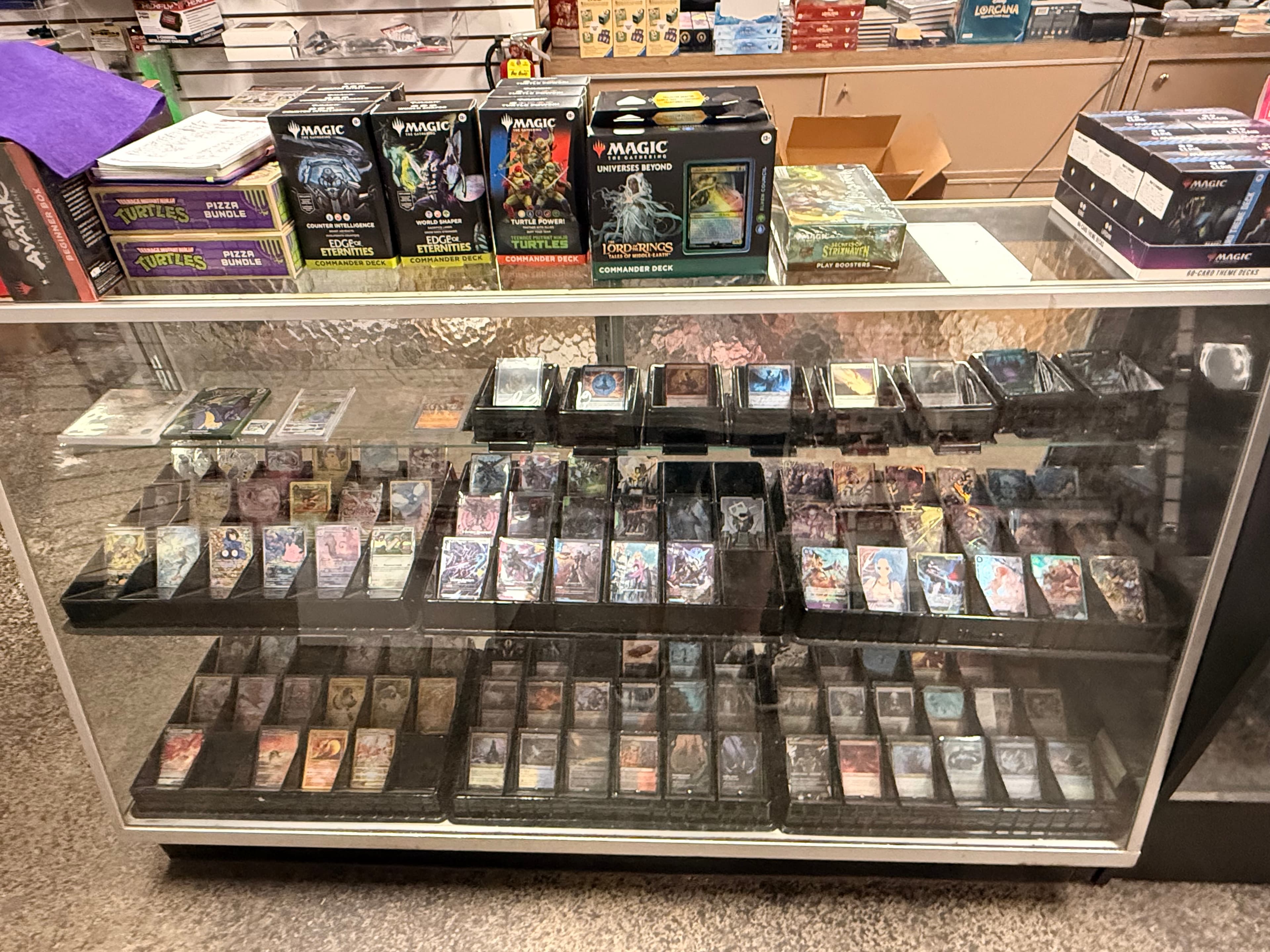 Glass display case showcasing Magic: The Gathering Commander decks and rows of individual trading cards.