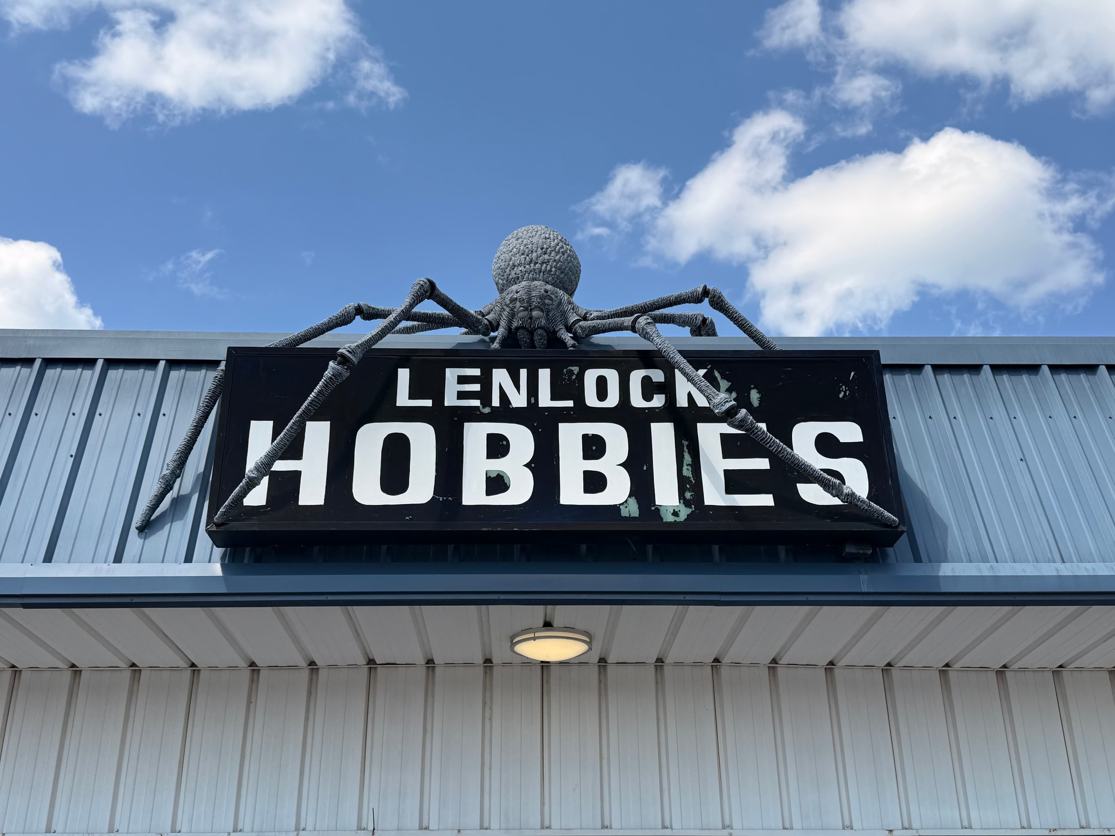 Large grey spider sculpture perched on a black Lenlock Hobbies sign against a blue sky.